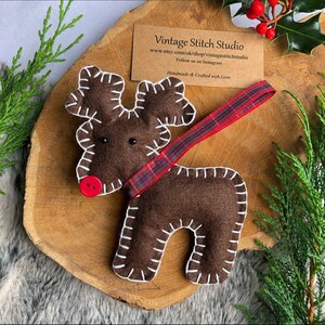 May include: A brown felt reindeer ornament with white stitching and a red button nose. The reindeer is wearing a red and black plaid ribbon. The ornament is handmade and says "Vintage Stitch Studio" on a card behind it.