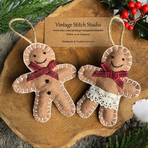 May include: Two felt gingerbread people ornaments with white stitching. One gingerbread person has a red and black plaid bow and the other has a white lace trim. Both have black button eyes and a smiling face. The ornaments are handmade and crafted with love.  www.etsy.com/uk/shop/vintagestitchstudio Follow us on Instagram