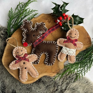 May include: Three felt Christmas ornaments: a brown reindeer with red and black plaid ribbon, a gingerbread person with red and black plaid ribbon, and a gingerbread person with white lace trim and a red and black plaid ribbon.
