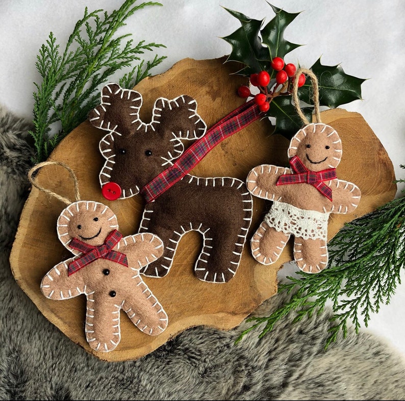 May include: Three felt Christmas ornaments: a brown reindeer with red and black plaid ribbon, a gingerbread person with red and black plaid ribbon, and a gingerbread person with white lace trim and a red and black plaid ribbon.