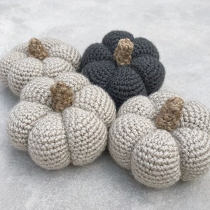 May include: Three crocheted pumpkins in different colors: beige, gray, and white. The pumpkins are all the same size and have a similar shape.