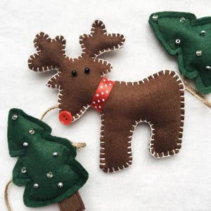 May include: A brown felt reindeer ornament with a red and white polka dot ribbon around its neck. The reindeer has a red button nose and black button eyes. There are two green felt Christmas tree ornaments with white beads on them.