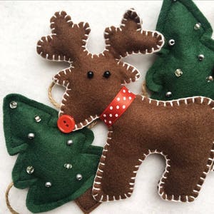 May include: A brown felt reindeer with white stitching and a red and white polka dot ribbon around its neck. The reindeer has black eyes and a red button nose. There are two green felt Christmas trees with white stitching and silver beads on them.