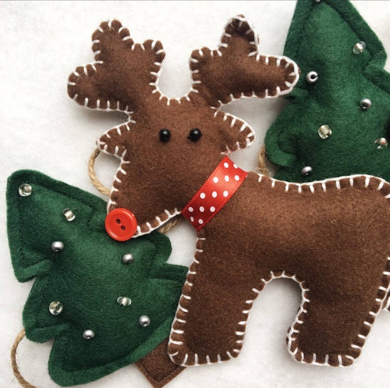 May include: A brown felt reindeer with white stitching and a red and white polka dot ribbon around its neck. The reindeer has black eyes and a red button nose. There are two green felt Christmas trees with white stitching and silver beads on them.