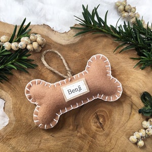 May include: A brown felt dog bone ornament with white stitching. The ornament has a small tag with the name "Benji" written on it.