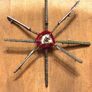 May include: A handcrafted starburst sculpture made from metal rods and wrapped with red and natural-colored yarn. The starburst is centered with a woven yarn design and has multiple metal rods extending outwards.