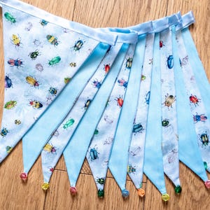 May include: A blue and white fabric bunting with a pattern of colorful beetles. The bunting is strung together with a blue ribbon and has colorful beads at the ends.