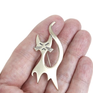 May include: A gold-toned cat brooch with a stylized design. The cat has pointed ears, a stern expression, and a long, curved tail. The brooch is held in a hand, showcasing its size and detail. The pin is a unique accessory.