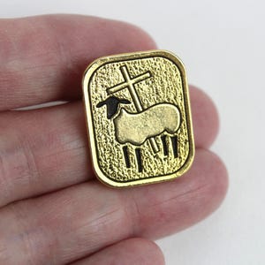 May include: A gold-colored rectangular pin featuring a lamb with a cross. The lamb is in the center, with a cross behind it. The pin has a textured background and a black outline.