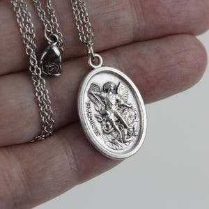 May include: Silver chain necklace with a silver oval pendant featuring a depiction of Saint Michael the Archangel. The pendant has the words "St. Michael" inscribed on it.