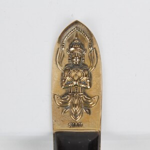May include: A gold-colored metal knife handle with a detailed design of a figure in a seated pose with hands clasped in prayer. The figure is surrounded by floral motifs. The handle is marked "Siam".