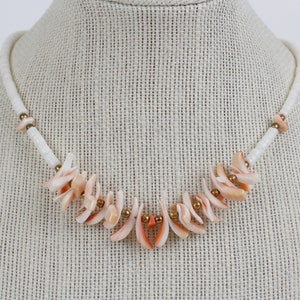 May include: A necklace featuring a white beaded strand with coral-colored shell accents. The necklace has a central design of shell fragments and small gold-colored beads. The necklace is displayed on a mannequin.