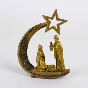 May include: A gold-colored nativity scene figurine. The scene includes figures of Mary, Joseph, and the baby Jesus in a manger. A star is above the figures, and the base has a textured, glittery finish. The figures are in a classic pose.
