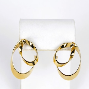 May include: A pair of gold-colored, oval-shaped earrings. Each earring features a double-loop design, with one loop intersecting the other. The earrings are displayed on a white stand against a white background. The earrings have a polished, reflective surface.