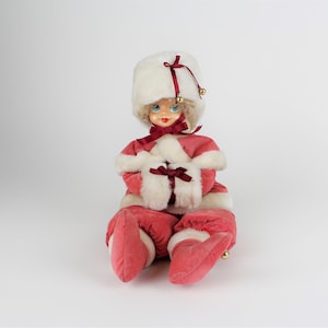 May include: A vintage Christmas doll wearing a pink velvet dress with white fur trim, a white fur hat with a red ribbon, and white fur mittens with a red ribbon. The doll has blue eyes and blonde hair.