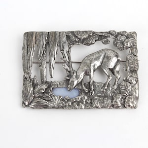 May include: A rectangular silver-toned brooch featuring a detailed relief of a deer drinking from a pool of water, set within a forest scene. The brooch has a pin closure on the back and is surrounded by a textured border of foliage and trees.