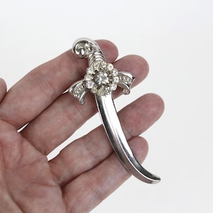 May include: A silver-toned brooch in the shape of a curved dagger. The hilt features a floral design with clear rhinestones. The blade is smooth and curved. The brooch is held in a hand against a white background.