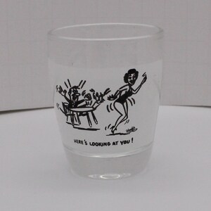 May include: A clear glass shot glass with a black and white cartoon illustration of a woman walking away from a table with three people sitting at it. The text "Here's looking at you!" is printed on the bottom of the glass.