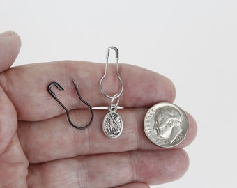 May include: A small, black safety pin, a silver safety pin with a religious pendant, and a dime are displayed. The dime shows a profile of a person. The items are on a light background.