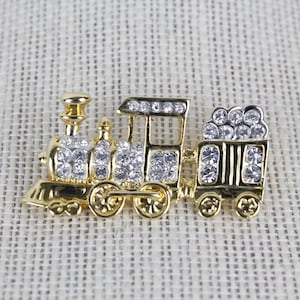 May include: A gold-toned train brooch with clear crystal accents. The locomotive and attached car are detailed with sparkling crystals. The brooch is set against a neutral, textured background, highlighting the jewelry's design.