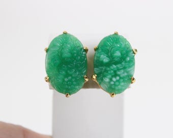 May include: A pair of oval-shaped earrings featuring carved green stone with floral motifs. Each piece is set in a gold-toned metal frame with four prongs securing the stone. The design displays intricate leaf and flower patterns on the surface.