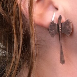 May include: A pair of copper axe earrings with a hammered finish. The earrings are hanging from the earlobe and are a unique and stylish accessory.