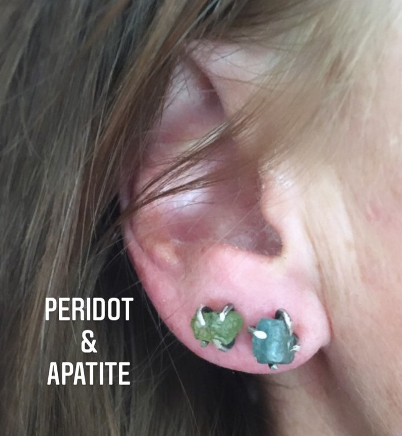 Pu&ograve; includere: Primo piano di un orecchio con due piccoli orecchini. L'orecchino sinistro ha una pietra di peridoto verde e l'orecchino destro ha una pietra di apatite blu. Gli orecchini sono incastonati in argento.