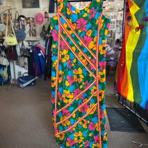 May include: A sleeveless knee-length dress featuring a vibrant floral pattern in shades of green, yellow, pink, and blue. The garment is accented with diagonal orange and white striped trim detailing across the front, creating a structured design.