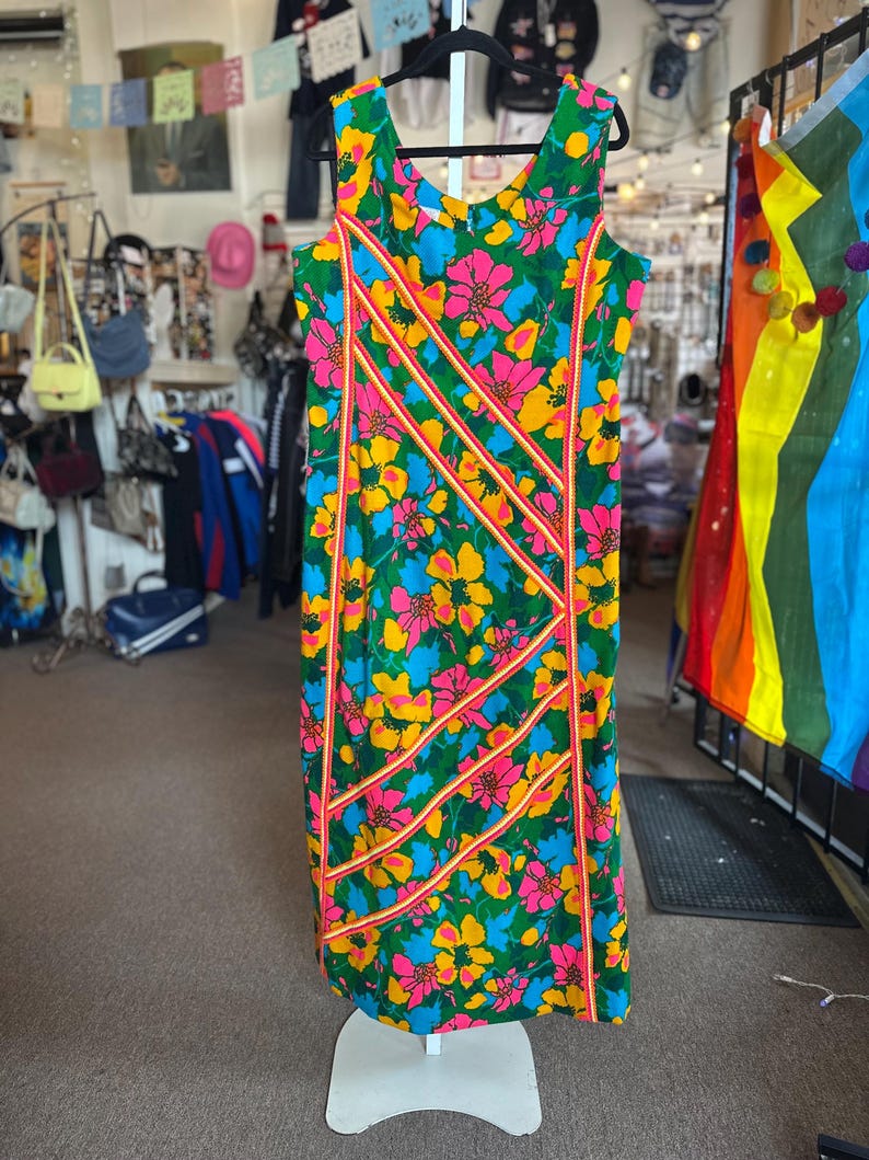 May include: A sleeveless knee-length dress featuring a vibrant floral pattern in shades of green, yellow, pink, and blue. The garment is accented with diagonal orange and white striped trim detailing across the front, creating a structured design.