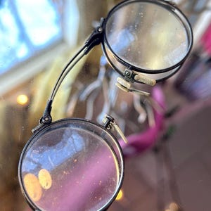 Puede incluir: Gafas redondas antiguas con montura negra y lentes transparentes. Las gafas tienen un diseño plegable con un asa decorativa y un puente. Las lentes están engastadas en una montura metálica con una bisagra.