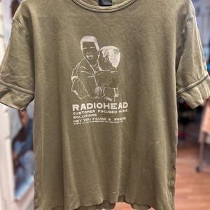May include: Olive green Radiohead t-shirt featuring a white graphic of two figures embracing. The text "RADIOHEAD" is above the graphic, with additional text below reading "CUSTOMER FOCUSED MUSIC SOLUTIONS" and "HEY YOU FOUND A FRIEND."