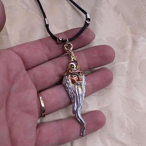 May include: A pendant necklace featuring a wizard design. The pendant has a gold-colored hat, a silver beard, and a face with a detailed expression. The necklace has a black cord with silver-colored accents.