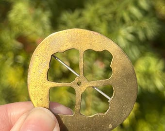 Pode incluir: Broche redondo, cor de latão, com um design de trevo de quatro folhas recortado no centro. Um alfinete prateado está preso na parte de trás. O broche é segurado em uma mão contra um fundo verde desfocado.
