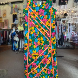 May include: A sleeveless dress featuring a vibrant floral pattern in shades of green, pink, yellow, and blue. The garment is accented with diagonal stripes of orange and white trim that run across the front, creating a structured geometric design.