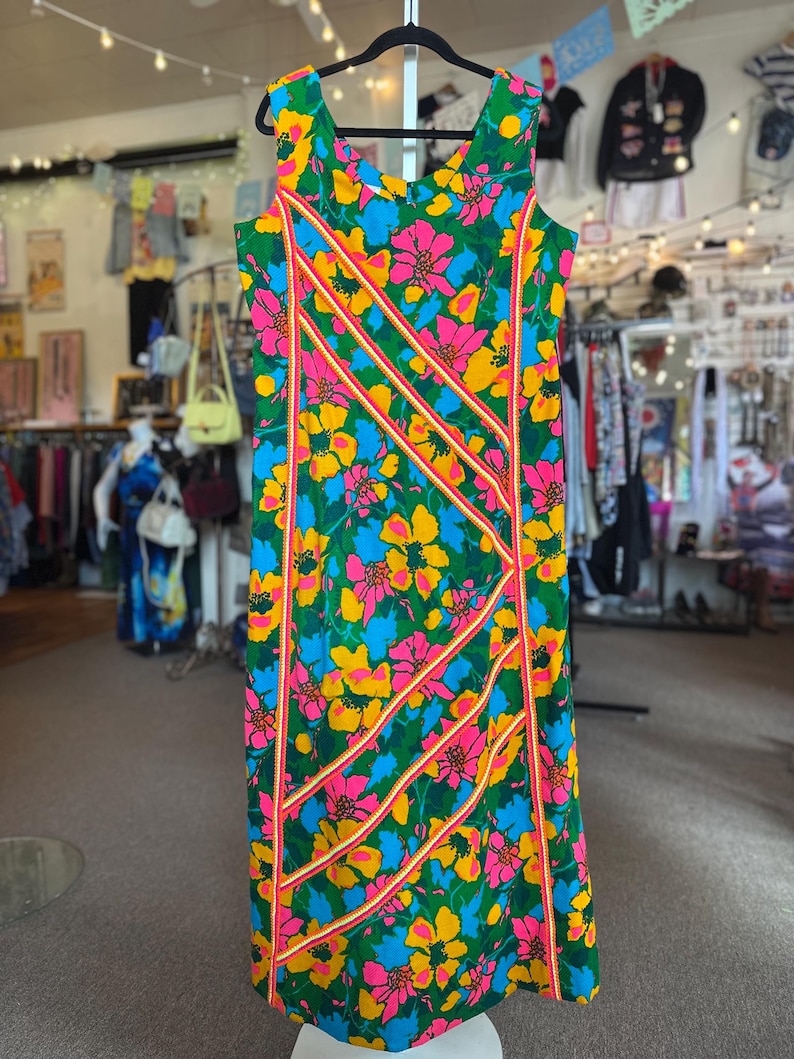 May include: A sleeveless dress featuring a vibrant floral pattern in shades of green, pink, yellow, and blue. The garment is accented with diagonal stripes of orange and white trim that run across the front, creating a structured geometric design.