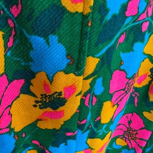 May include: A textile fabric featuring a vibrant floral pattern with large, bold flowers in shades of hot pink, bright yellow, and sky blue set against a deep forest green background. The material has a visible woven texture and a vertical seam.
