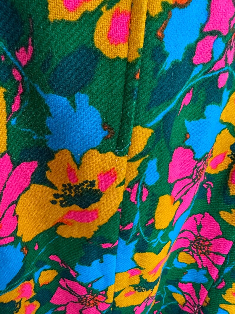 May include: A textile fabric featuring a vibrant floral pattern with large, bold flowers in shades of hot pink, bright yellow, and sky blue set against a deep forest green background. The material has a visible woven texture and a vertical seam.