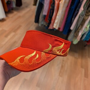 Puede incluir: Una visera roja con un dise&ntilde;o de llamas doradas. La visera est&aacute; hecha de un material suave y c&oacute;modo y tiene una visera curva.