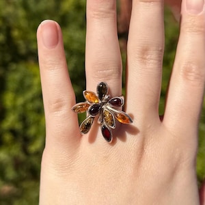 Puede incluir: Un anillo de plata con un diseño floral con piedras de ámbar. Los pétalos del anillo muestran una variedad de tonos ámbar, incluyendo tonos de naranja, marrón y verde. El anillo se encuentra sobre un fondo verde borroso.