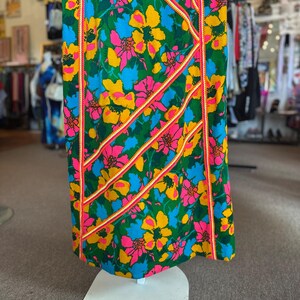May include: A floor-length dress featuring a vibrant floral pattern in shades of green, yellow, blue, and pink. The garment is accented with diagonal and vertical strips of orange and white braided trim that run across the fabric surface.