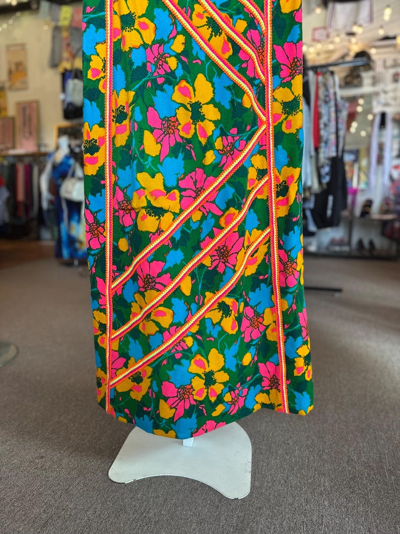 May include: A floor-length dress featuring a vibrant floral pattern in shades of green, yellow, blue, and pink. The garment is accented with diagonal and vertical strips of orange and white braided trim that run across the fabric surface.