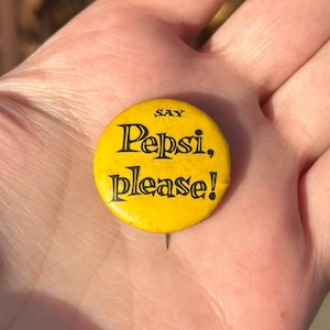 May include: A round, yellow Pepsi button with the text "Say Pepsi, please!" in black lettering. The button has a pin on the back and is held in a hand.