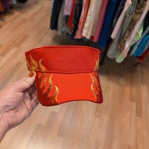 Puede incluir: Una visera roja con bordado de llamas doradas.