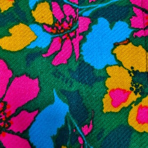 May include: A textile fabric featuring a vibrant floral pattern with large, stylised flowers in shades of hot pink, bright yellow, and sky blue set against a deep forest green background. The material has a visible woven texture throughout.