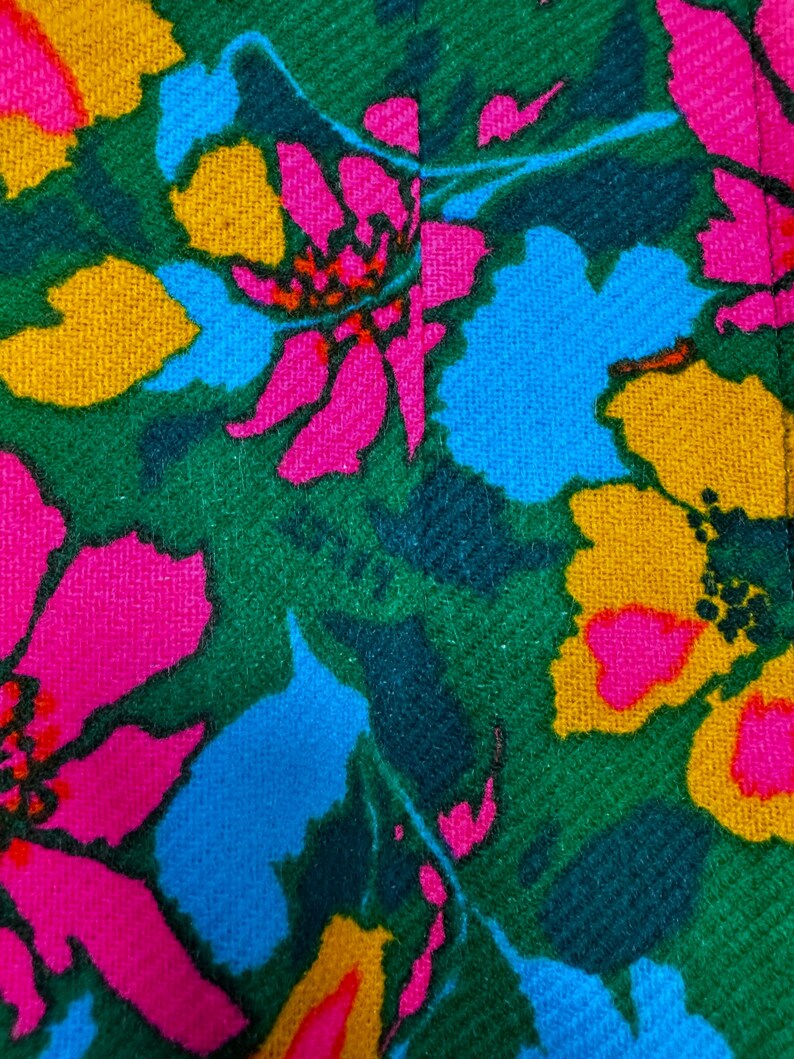 May include: A textile fabric featuring a vibrant floral pattern with large, stylised flowers in shades of hot pink, bright yellow, and sky blue set against a deep forest green background. The material has a visible woven texture throughout.