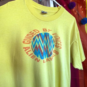 May include: A yellow t-shirt with the text "GUIDED BY VOICES ALIEN LANES" in orange. The design includes a circular graphic with blue, green, and red zigzags. The shirt is hanging on a white hanger.