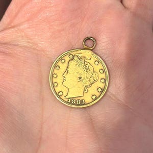 May include: A gold-colored coin pendant with a loop for a chain. The coin features a profile of a woman, the word "LIBERTY", and the year "1883". The pendant is round with star-shaped accents around the edge.
