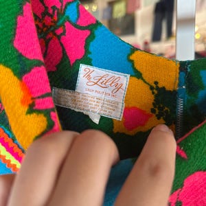 May include: A sleeveless dress featuring a vibrant floral pattern in neon pink, bright blue, and yellow on a green background. A white label is sewn at the neckline with the text The Lilly Lilly Pulitzer Inc. and care instructions.