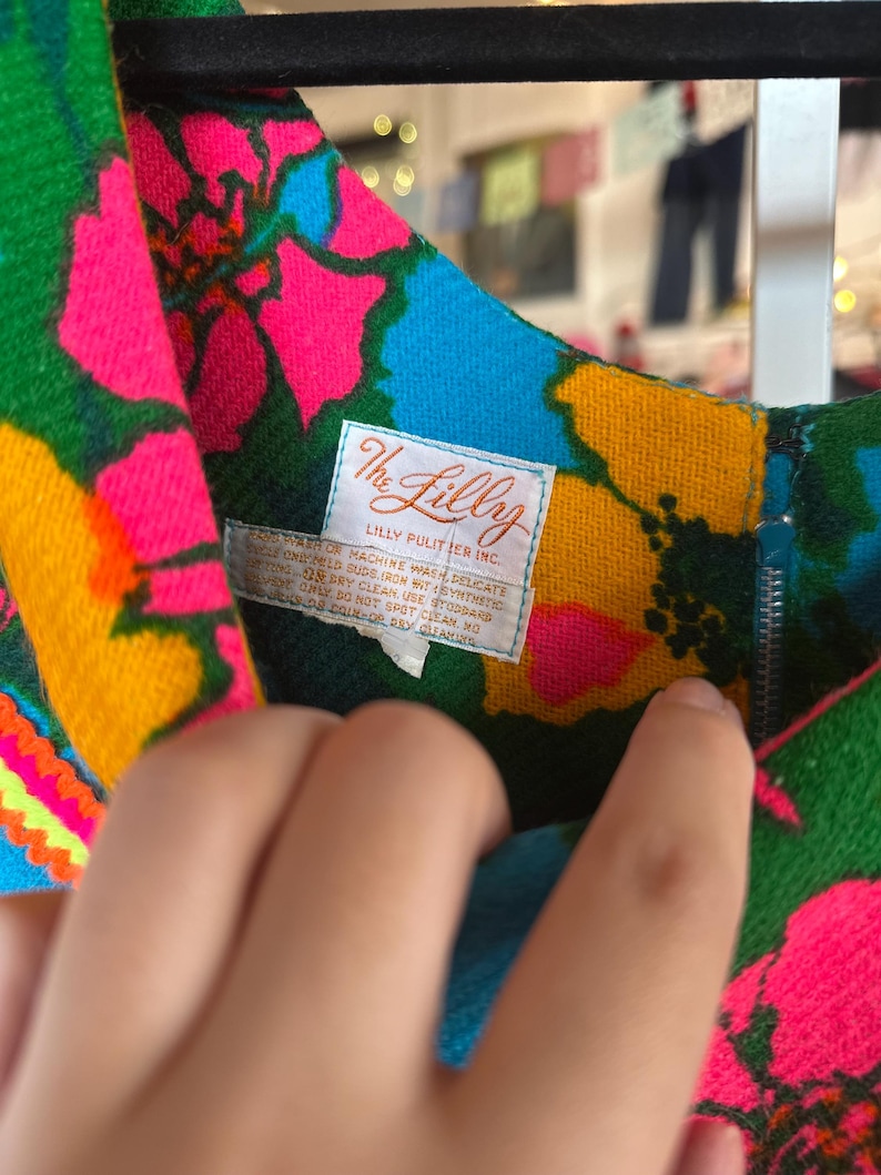 May include: A sleeveless dress featuring a vibrant floral pattern in neon pink, bright blue, and yellow on a green background. A white label is sewn at the neckline with the text The Lilly Lilly Pulitzer Inc. and care instructions.