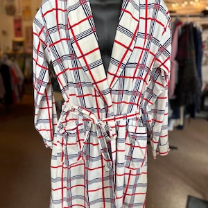 May include: A white robe featuring a plaid pattern in red and navy blue lines. The garment has long sleeves, a shawl collar, two front patch pockets, and a matching fabric tie belt at the waist to secure the front closure of the robe.